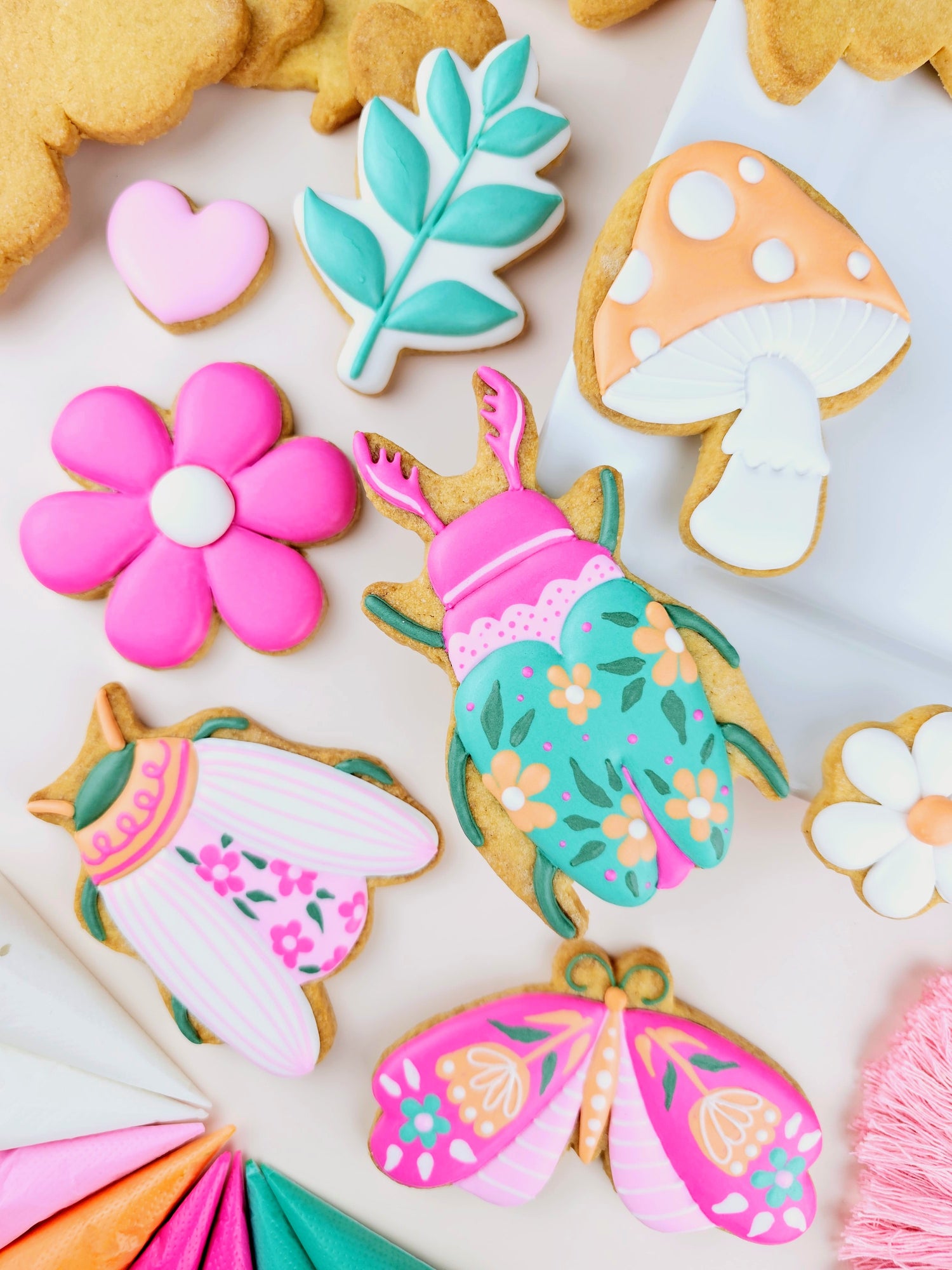 A styled flat-lay of decorated sugar cookies shaped like a leaf, mushroom, flower, butterfly, and dress, arranged on a white surface with colorful piping bags partially visible at the bottom left.

Cookie decorating Workshop/ Class Cape Town